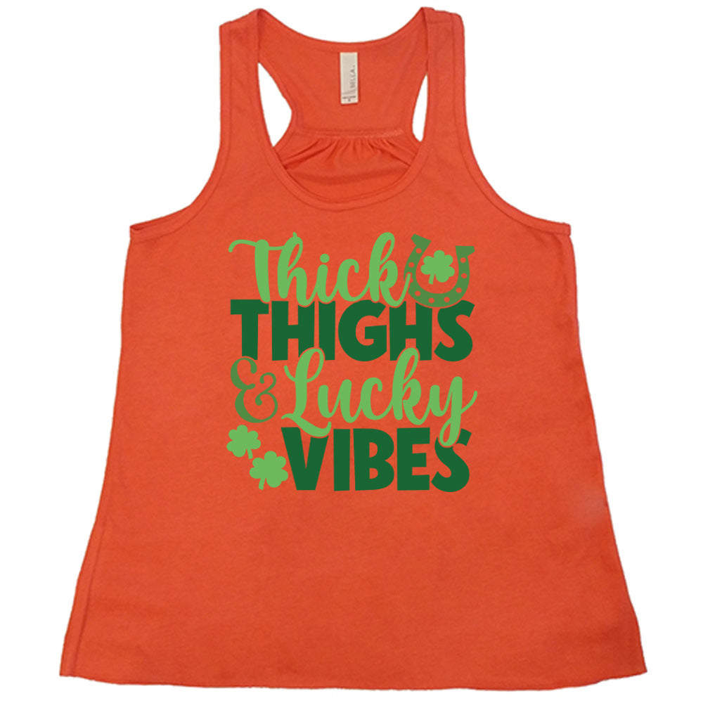 coral flowy tank with the words "thick thighs & lucky vibes" on it, with graphics of a horseshoe and a few shamrocks