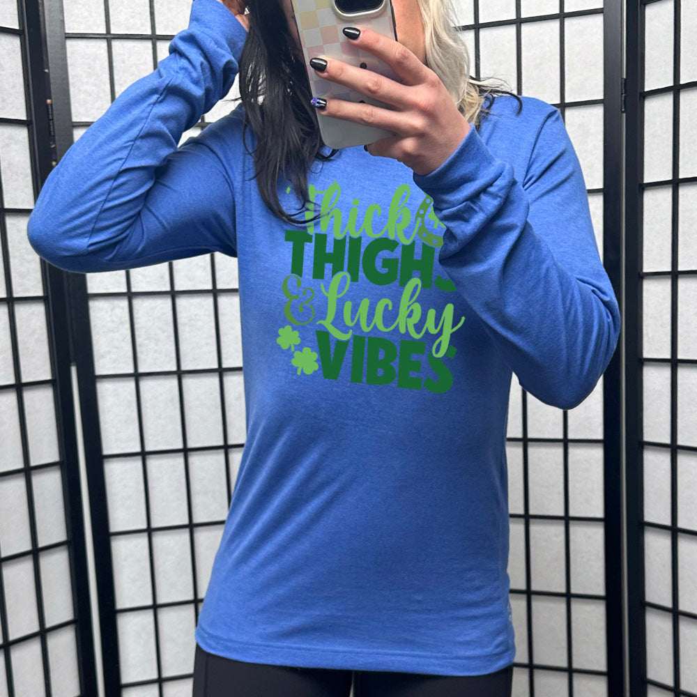 heather true royal long sleeve with the words "thick thighs & lucky vibes" on it, with graphics of a horseshoe and a few shamrocks