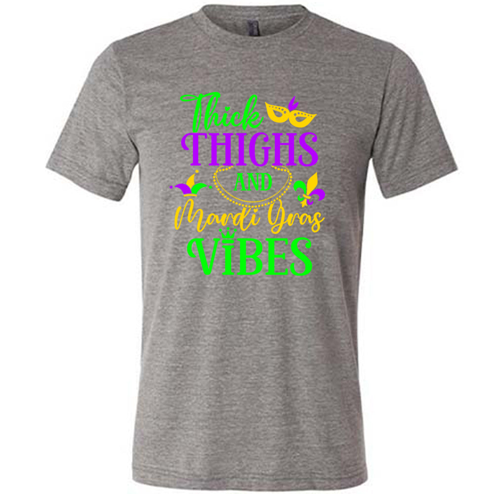 grey unisex tee with the words "thick thighs and mardi gras vibes" on it in varying font, with small graphics of mardi gras beads, fleur de lis, a joker hat and a mask