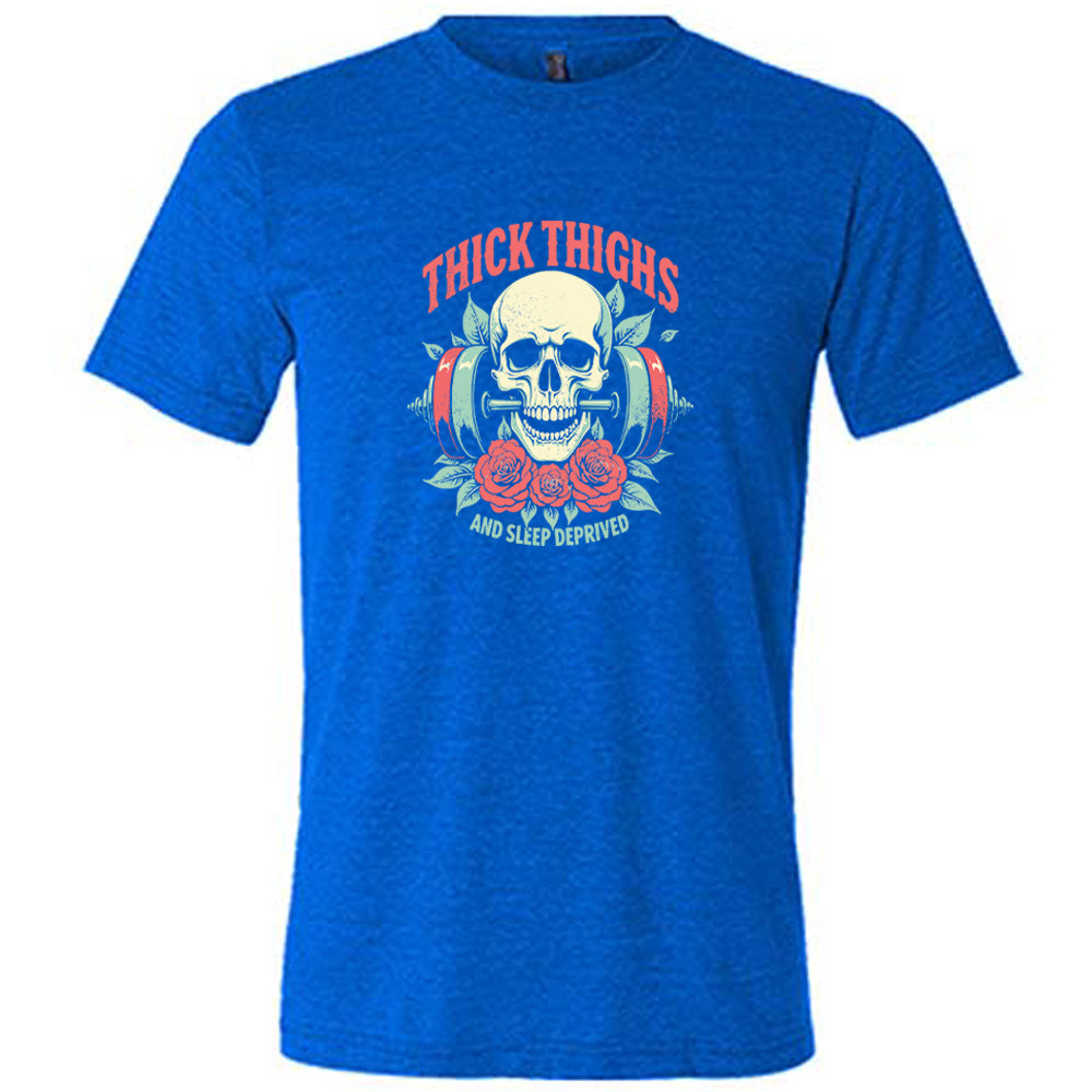 Blue colored shirt with a skull, flowers, and barbell graphic on it that says "Thick Thighs And Sleep Deprived" 