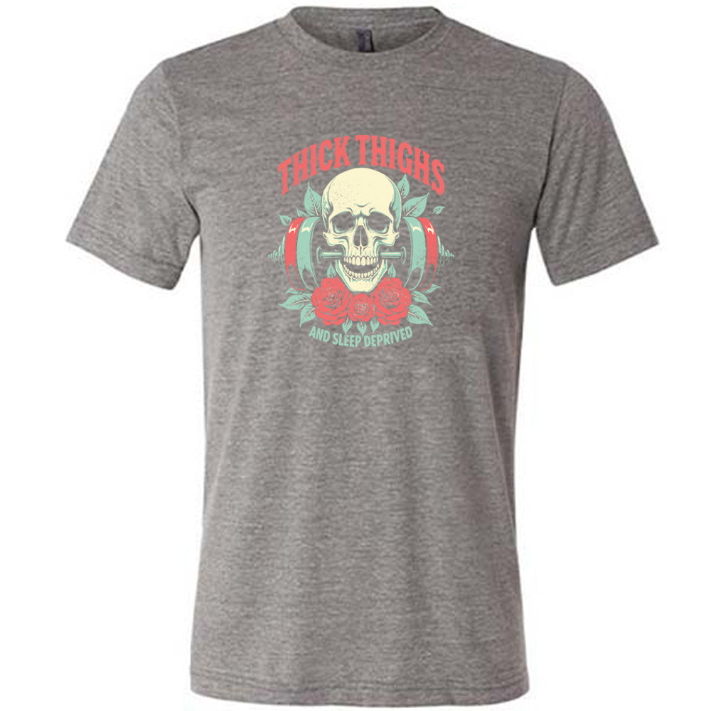 Grey colored shirt with a skull, flowers, and barbell graphic on it that says "Thick Thighs And Sleep Deprived" 