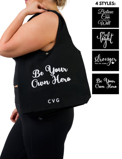 model holding the "Be Your Own Hero" black tote bag, 4 unique styles to choose from "Believe You Can And You Will", "Be The Light", "Stronger Than The Storm", and "Be Your Own Hero"