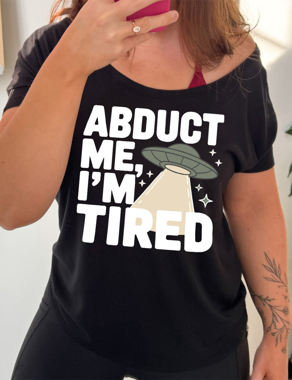 black slouchy tee with the words "abduct me, I'm tired" on it, with a graphic of an alien spaceship, with a beam extended, and a few little stars