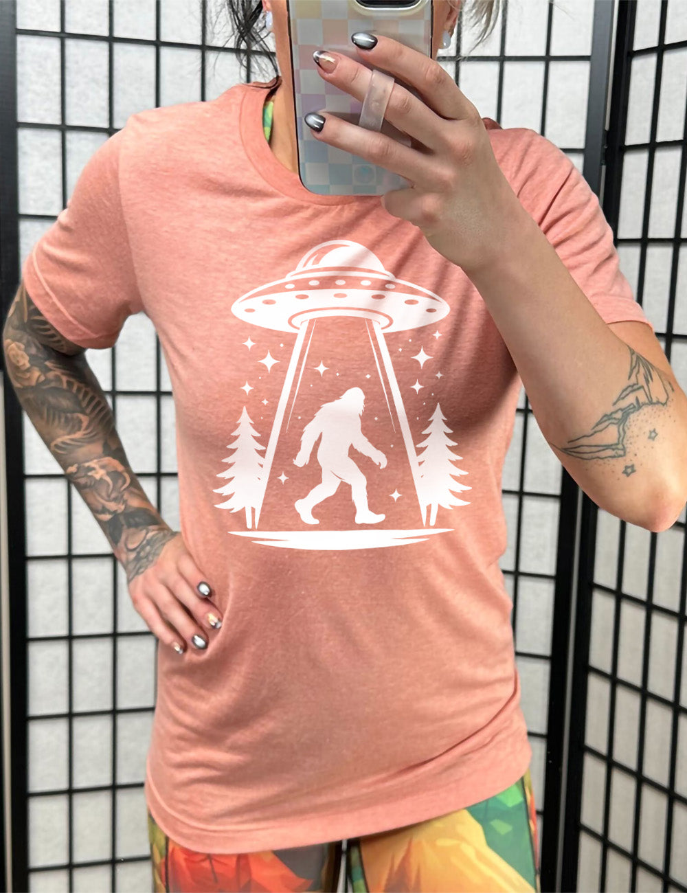 sunset unisex tee with the graphic of an alien spaceship beaming bigfoot up, with stars surrounding, and couple pine trees