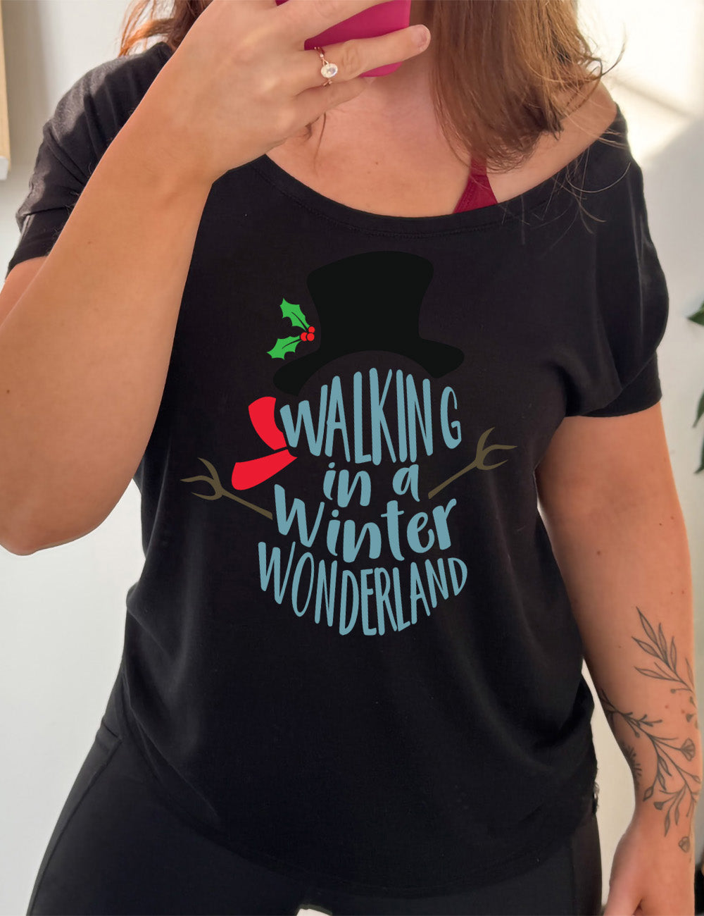 black slouchy tee with the words in the stacked shape of a snowman saying "walking in a winter wonderland" with the graphic completion of the snowman with stick arms, a red scarf, and a top hat with a holly branch accessory on it