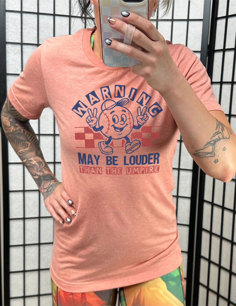 sunset unisex tee with the words "warning may be louder than the umpire" on it, with the graphic of baseball with a face, wearing a hat, holding up two peace signs, and walking along, with a checkered border behind it