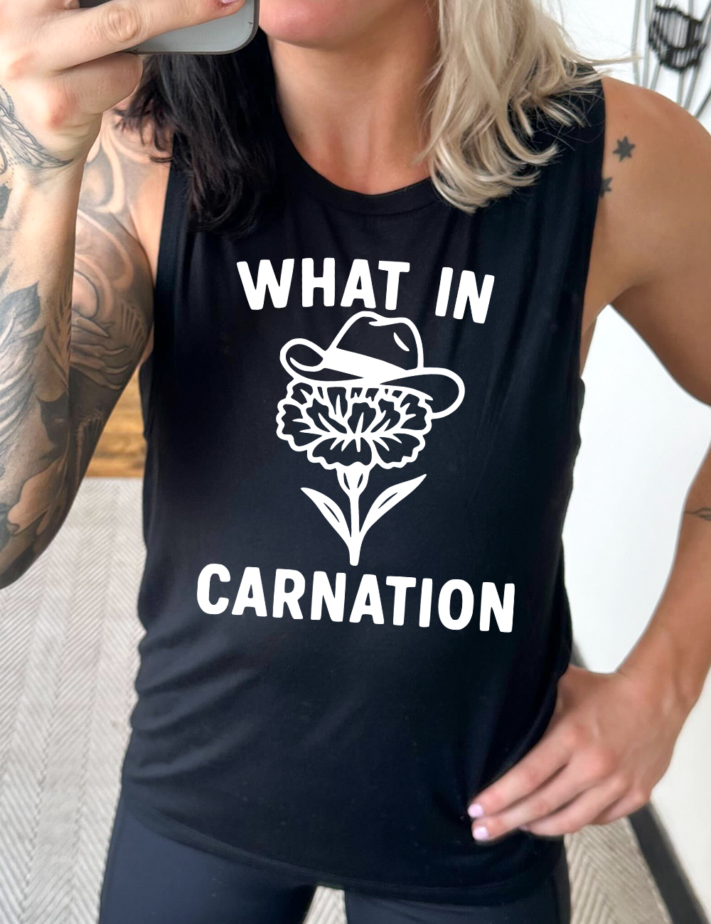 black muscle tank with the words "What in carnation" on it, around the graphic of a carnation, wearing a cowboy hat