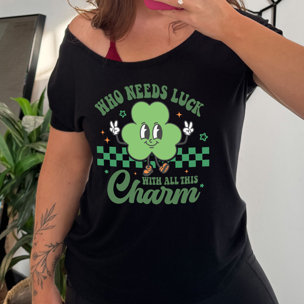 black slouchy tee with the words "who needs luck with all this charm" on it, with the graphic of a cheerful shamrock (with eyes, nose and smiling mouth) giving two peace signs with its hands, in front of a partial checkered background, with a few stars around