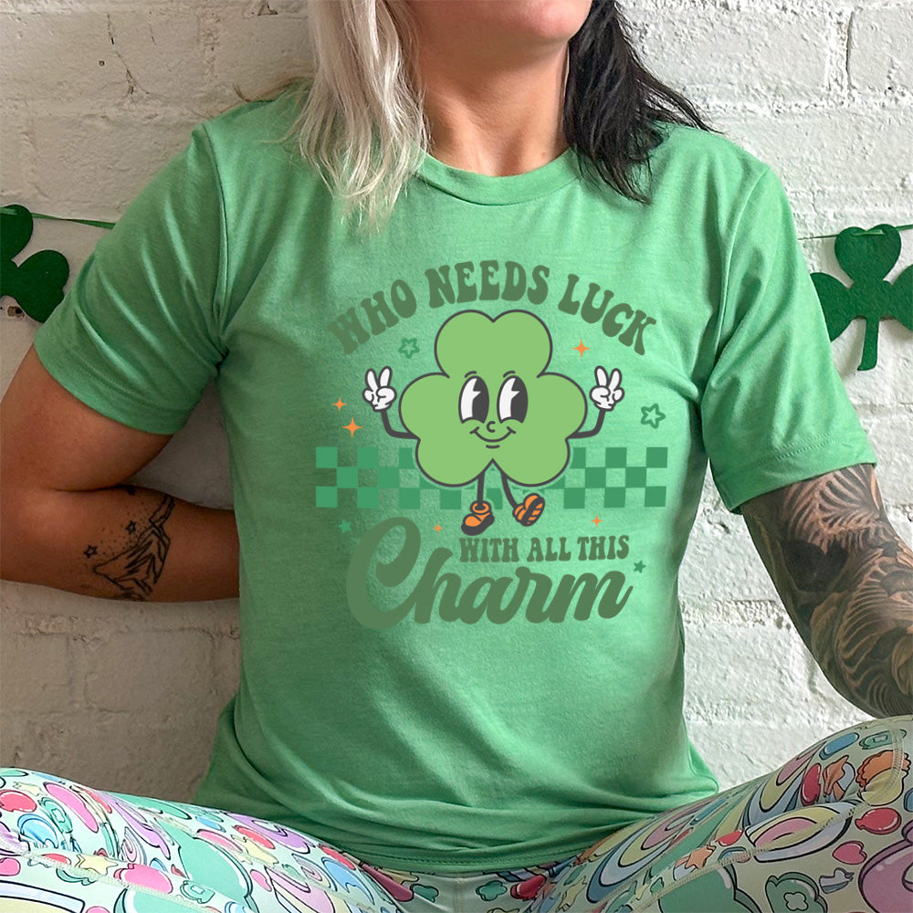 lucky green unisex tee with the words "who needs luck with all this charm" on it, with the graphic of a cheerful shamrock (with eyes, nose and smiling mouth) giving two peace signs with its hands, in front of a partial checkered background, with a few stars around