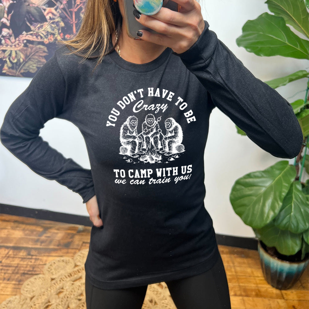 heather black long sleeve with the words "you don't have to be crazy to camp with us we can train you!" on it with the graphic of three sasquatches toasting marshmallows over a campfire