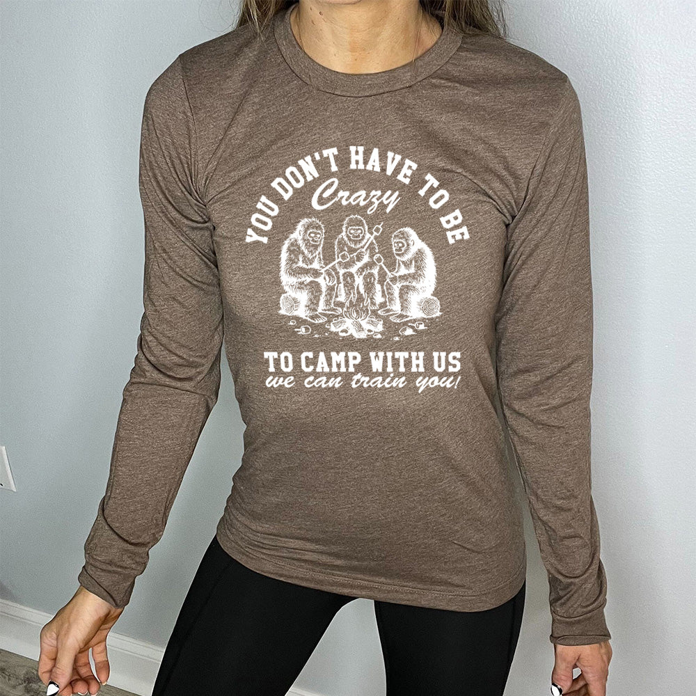 heather brown long sleeve with the words "you don't have to be crazy to camp with us we can train you!" on it with the graphic of three sasquatches toasting marshmallows over a campfire