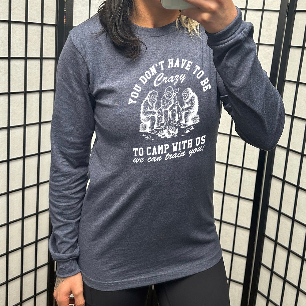 heather navy long sleeve with the words "you don't have to be crazy to camp with us we can train you!" on it with the graphic of three sasquatches   toasting marshmallows over a campfire