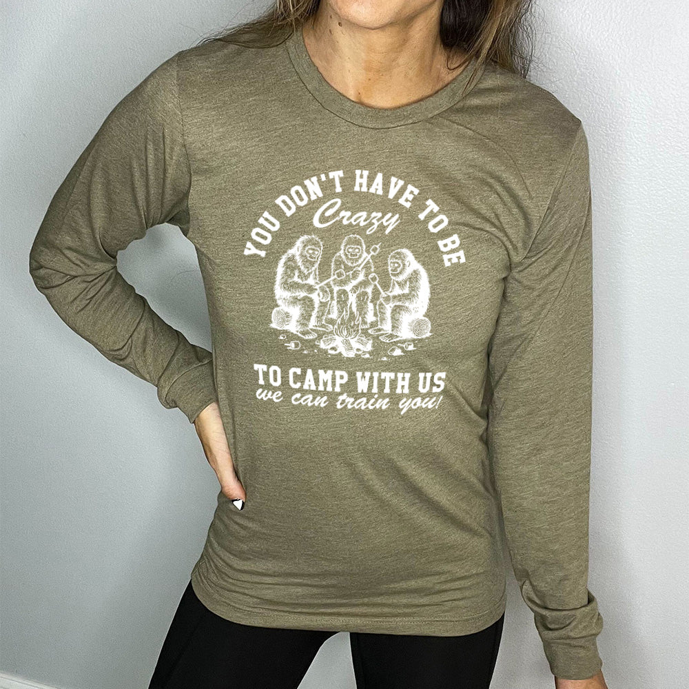 heather olive long sleeve with the words "you don't have to be crazy to camp with us we can train you!" on it with the graphic of three sasquatches  toasting marshmallows over a campfire
