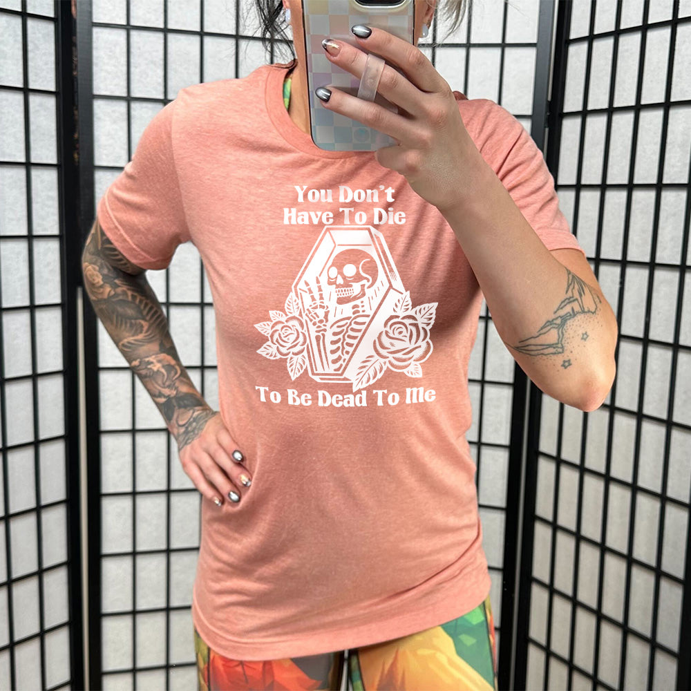 sunset unisex tee with the words "You don't have to die to be dead to me" on it with the graphic of a a skeleton in a coffin, giving the peace sign, with two flowers on either side of the coffin