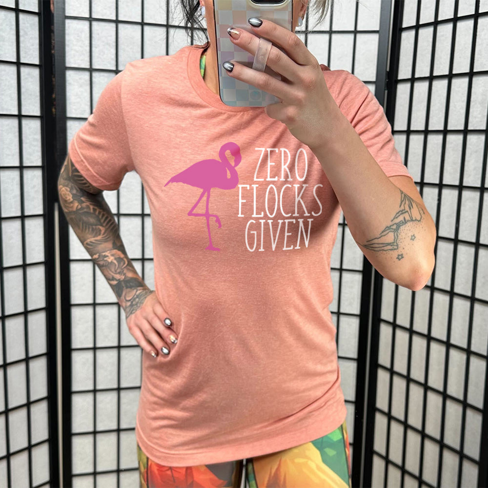 sunset unisex tee with the words "zero flocks given" on it, with the pink silhouette of a flamingo, leg raised, next to the words