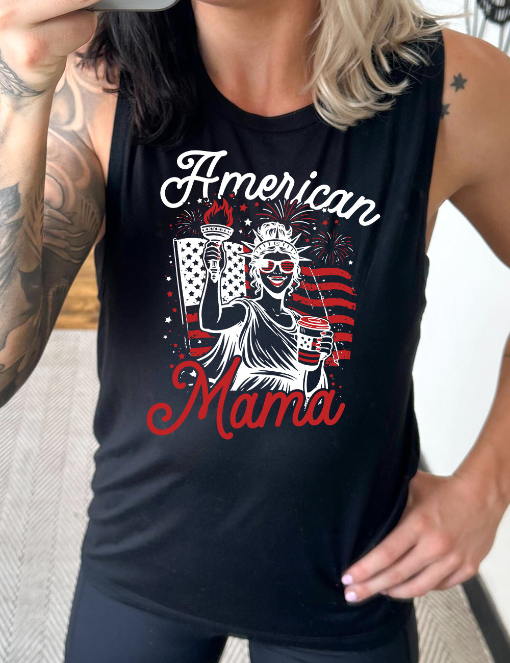 black muscle tank with the graphic of the statue of liberty in front of an American flag, fireworks above, wearing American flag sunglasses, holding a American flag coffee cup, with the words "American Mama" surrounding it
