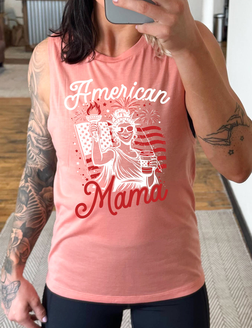 peach muscle tank with the graphic of the statue of liberty in front of an American flag, fireworks above, wearing American flag sunglasses, holding a American flag coffee cup, with the words "American Mama" surrounding it