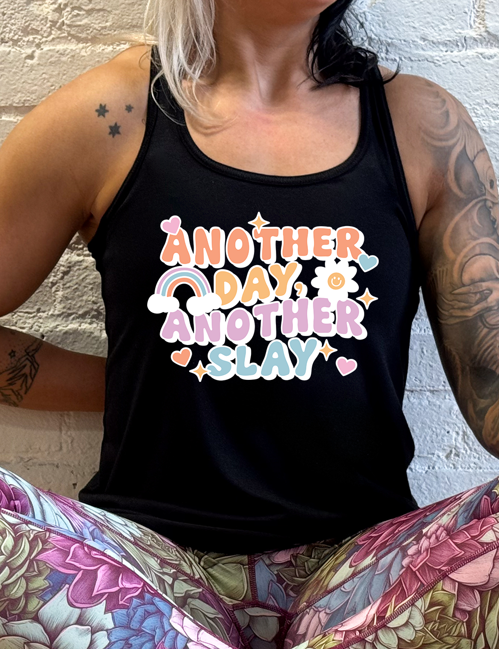 black racerback flowy tank with the words "another day, another slay" on it, with little graphics of stars, hearts, a sun, and a rainbow