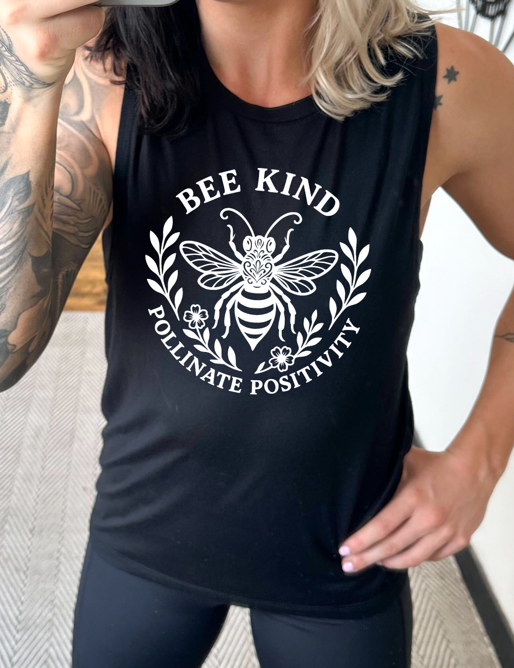 black muscle tank with the words "bee kind pollinate positivity" on it, arranged in two arcs, forming a circled effect, with two branches and two flowered branches within the circle, and a large fancy bee in the center