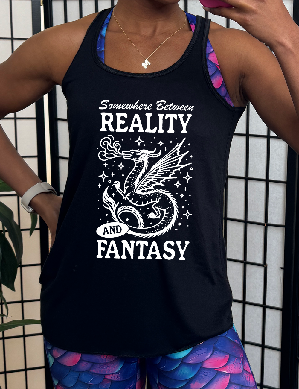 black racerback flowy tank with the words "somewhere between reality and fantasy" on it, around the graphic of a dragon among stars, breathing fire