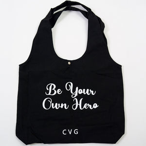 "be your own hero" saying on a black tote bag