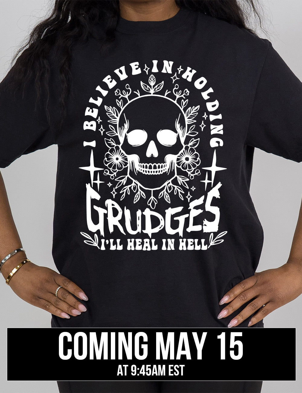 black modern tee with print placement on the front, with the graphic of a skull among flowers, and  words arching over it and words underneath, saying "I believe in holding GRUDGES I'll heal in hell", coming may 15th