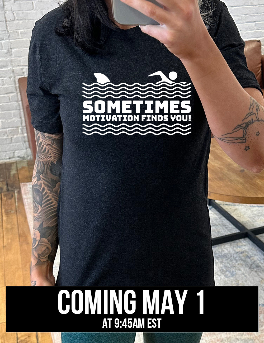 black unisex tee with the words "sometimes motivation finds you!" in between the lined graphic of waves of the sea, and on top of the waves is the graphic of a figure swimming away from a fin in the water behind, coming soon
