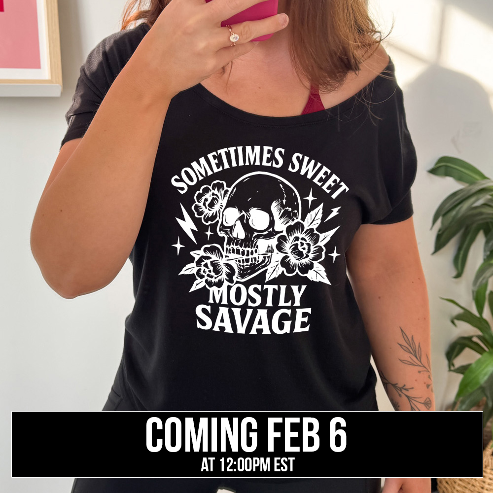 black slouchy tee with the words "sometimes sweet mostly savage on it with the graphic of a skull holding a flower between its teeth, with a couple flowers and a few stars and lightening bolts around it, coming soon