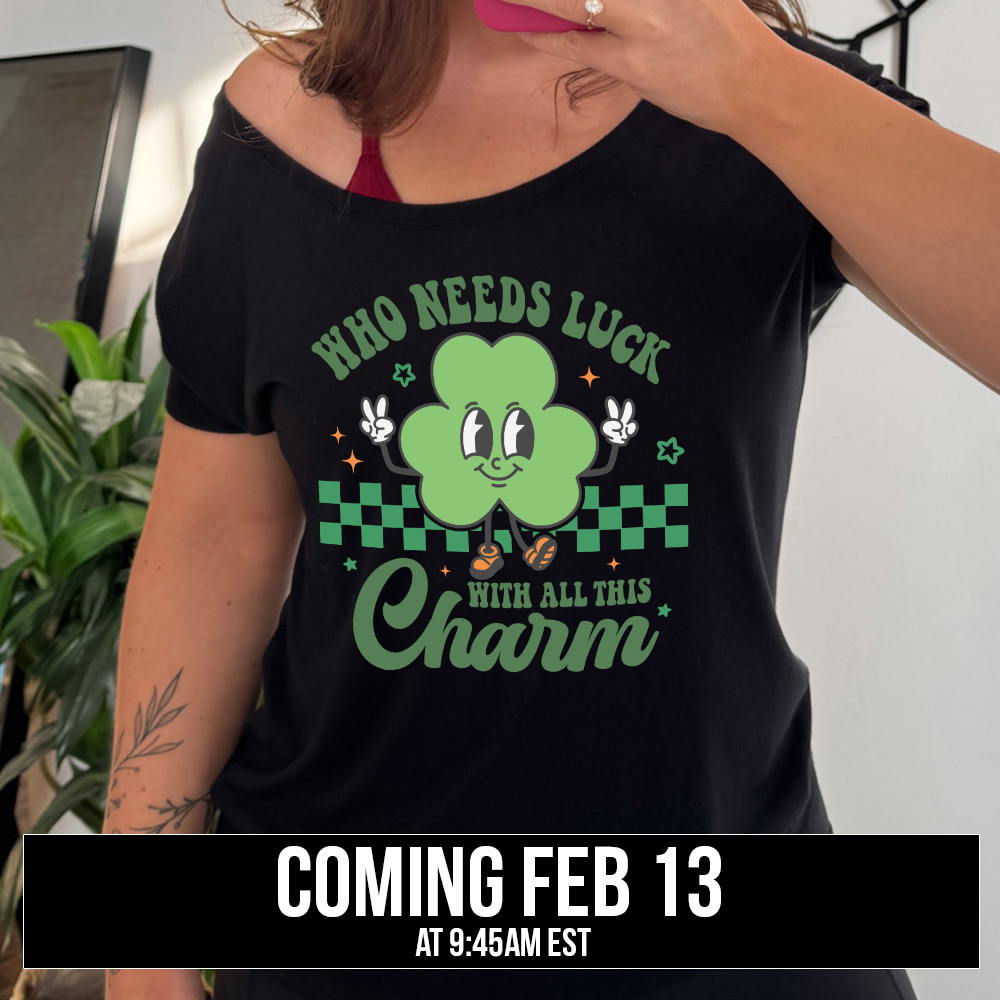black slouchy tee with the words "who needs luck with all this charm" on it, with the graphic of a cheerful shamrock (with eyes, nose and smiling mouth) giving two peace signs with its hands, in front of a partial checkered background, with a few stars around, coming soon