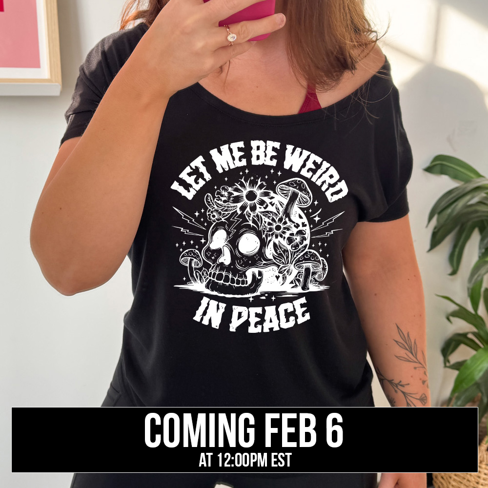 black slouchy tee with the words "let me be weird in peace" on it with the graphic of a skull on the ground with mushrooms growing on and around it, as well as flowers growing on it, surrounded by little stars and lightening bolts, coming soon