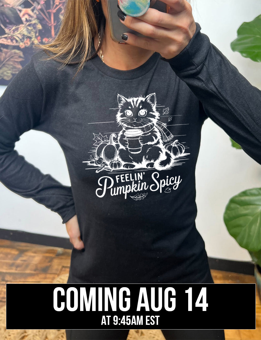 heather black long sleeve with the graphic of a cat, wearing a scarf, next to a few pumpkins of various sizes and a few falling leaves, holding a takeaway coffee, and the words underneath saying "feelin' pumpkin spicy" coming august 14th