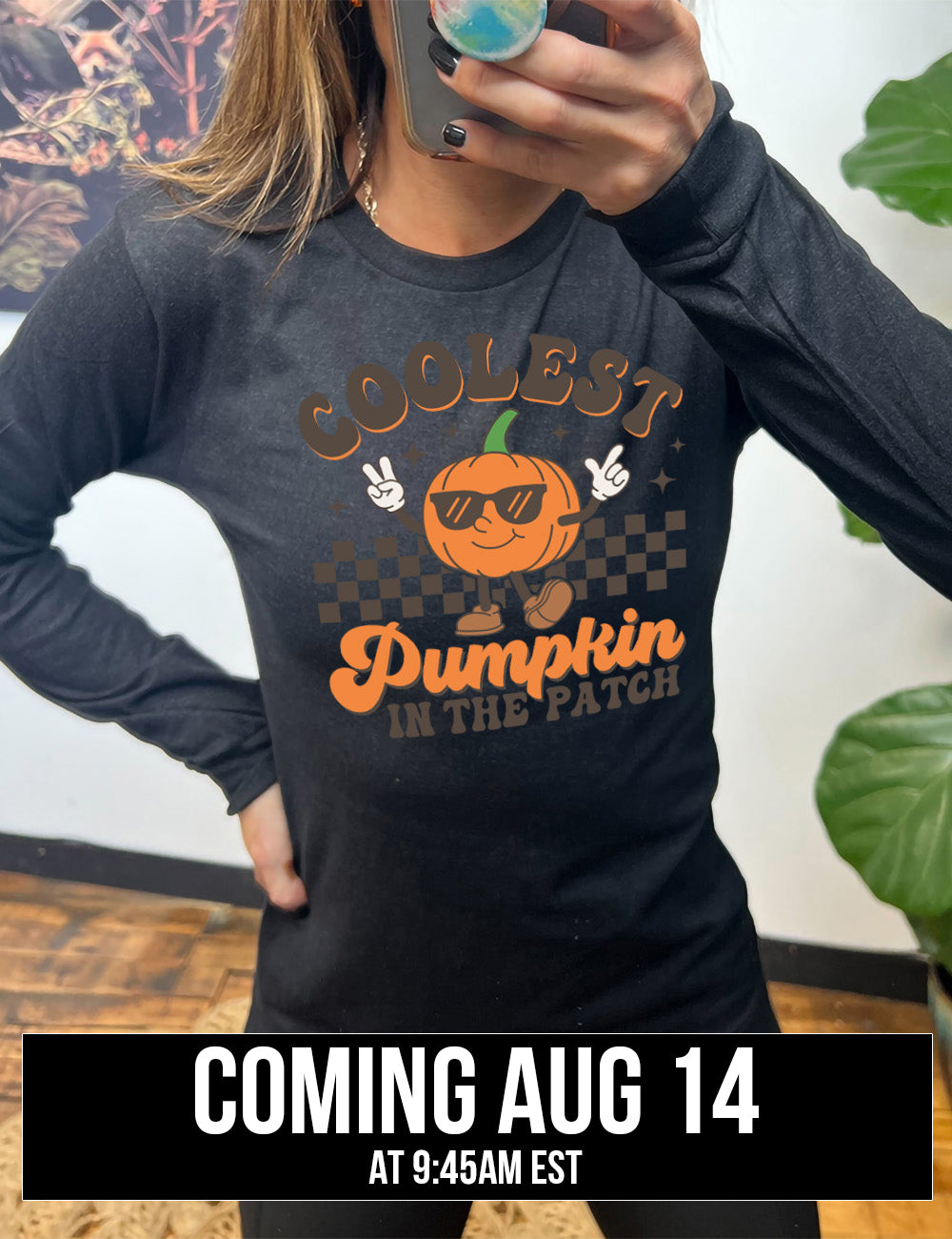 heather black long sleeve with the graphic of a pumpkin, wearing sunglasses, both arms raised, foot kicking out, in front of a checkered banner, surrounded by a few little stars, with the words around it saying "coolest pumpkin in the patch", coming august 14th