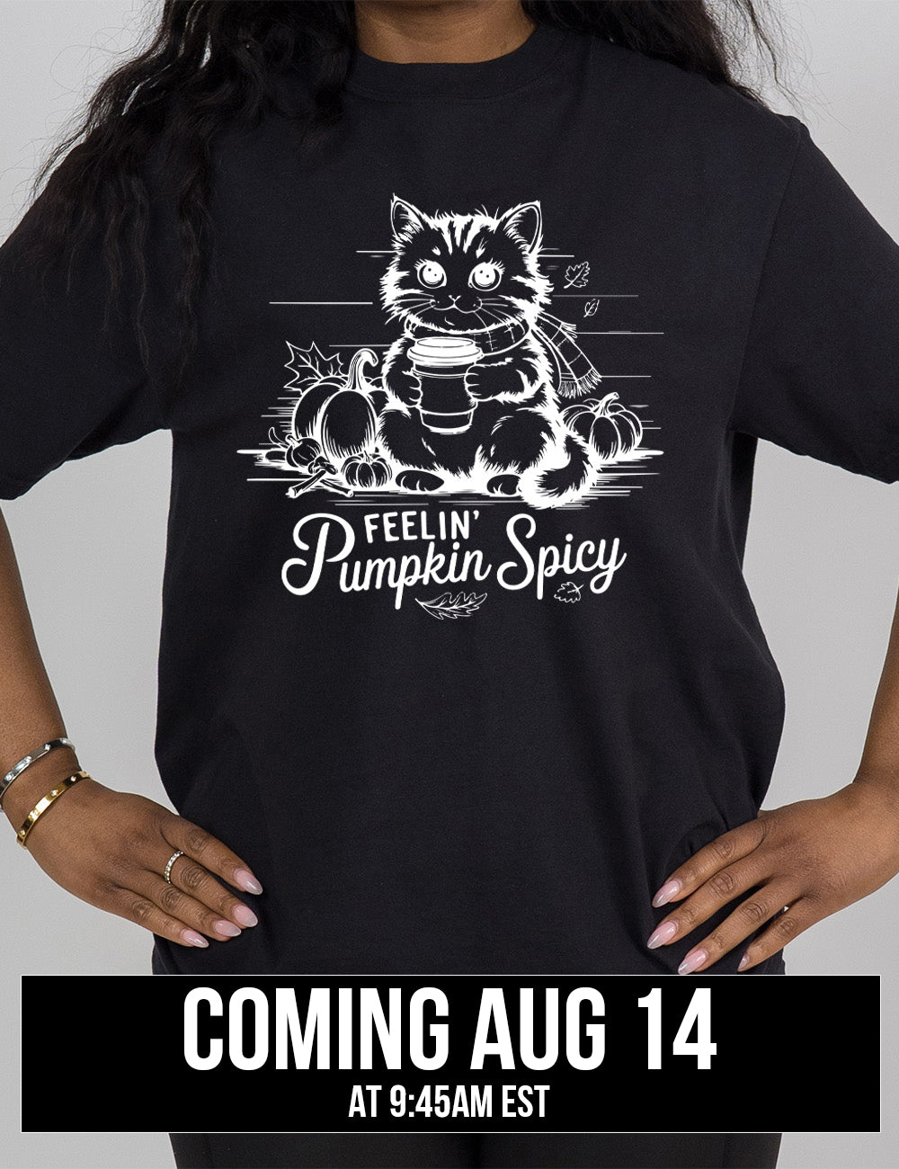 black modern tee with the graphic of a cat, wearing a scarf, next to a few pumpkins of various sizes and a few falling leaves, holding a takeaway coffee, and the words underneath saying "feelin' pumpkin spicy" coming august 14th