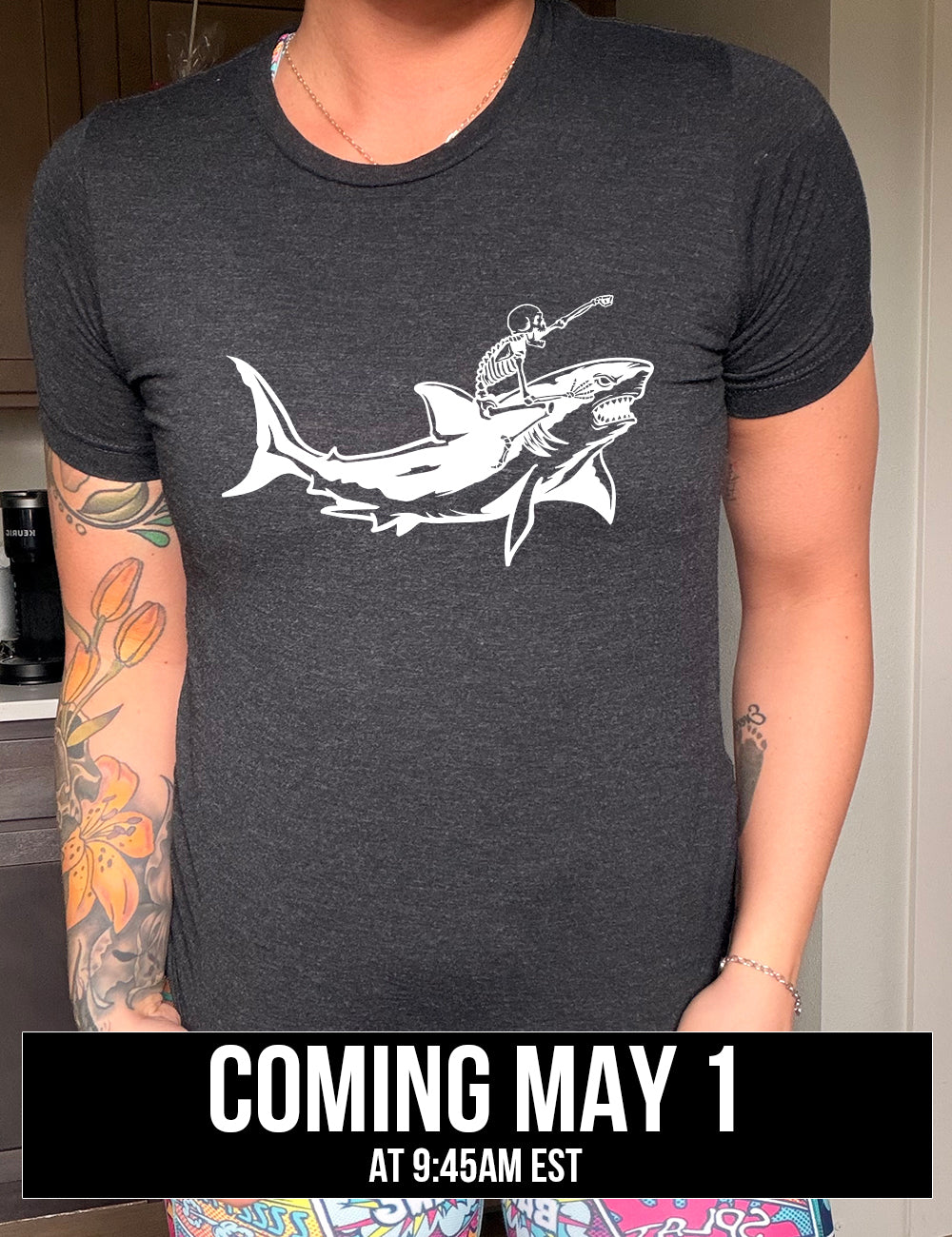 black unisex tee with the graphic of a skeleton, with its fist pointing onwards and its mouth open, riding a shark, who has a fierce expression, coming soon