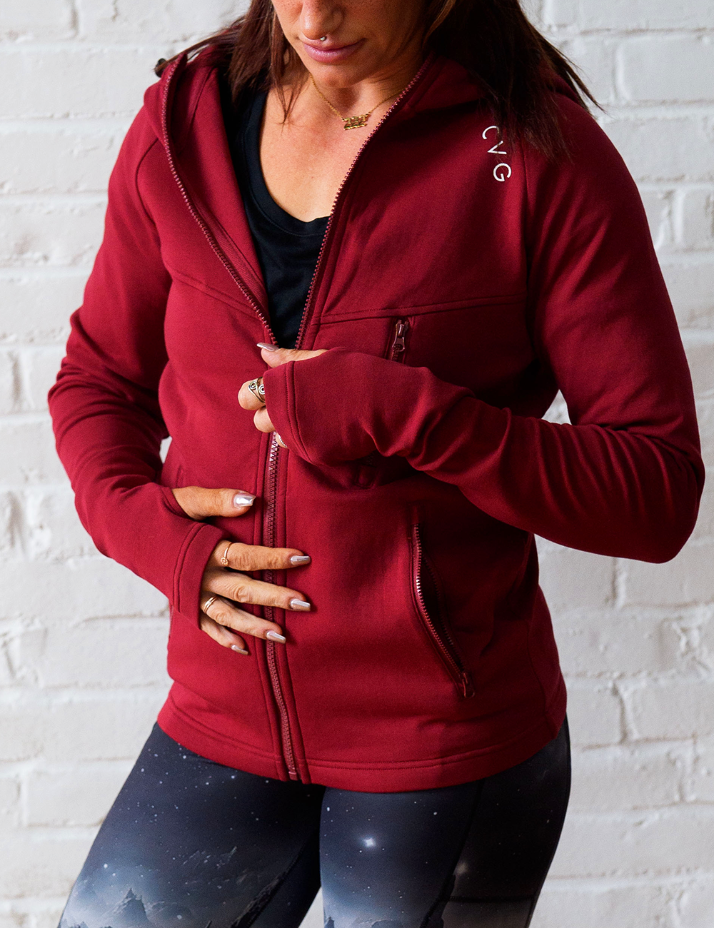 close up of the dark red rest day zip up being zipped up, the unknown  dark mountain with stars pattern leggings are also being worn against a white brick background