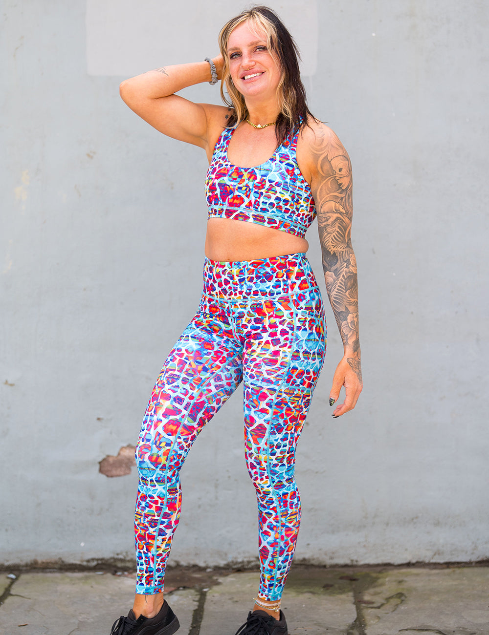 model wearing the turquoise, coral, pink, orange, purple, and yellow mosaic style pattern full length leggings and bra in front of a concrete wall