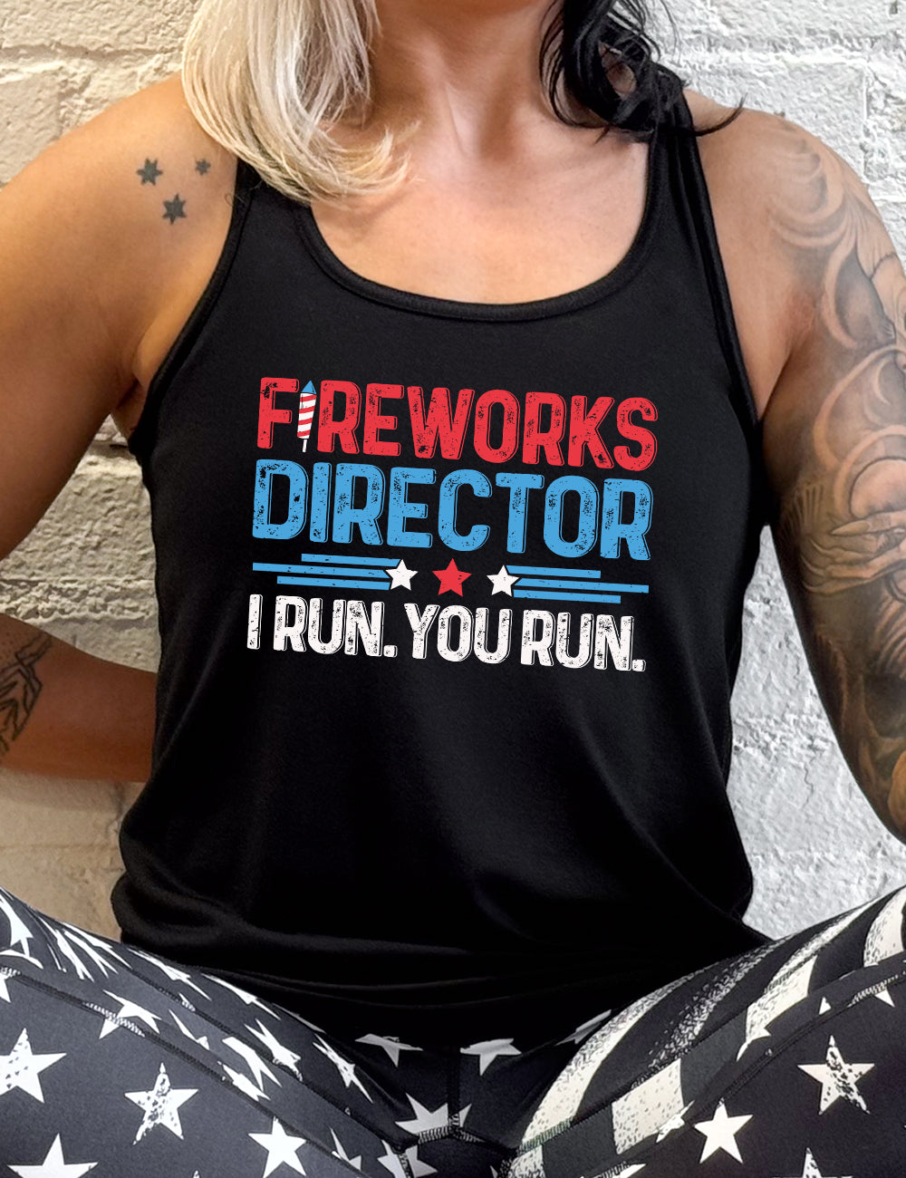 black flowy racerback tank with the words "Fireworks Director -- I run. You run." on it, with the "i" of "fireworks" in the shape of a firework, and some stripes with a few stars in between the word "director" and "I run. You run."