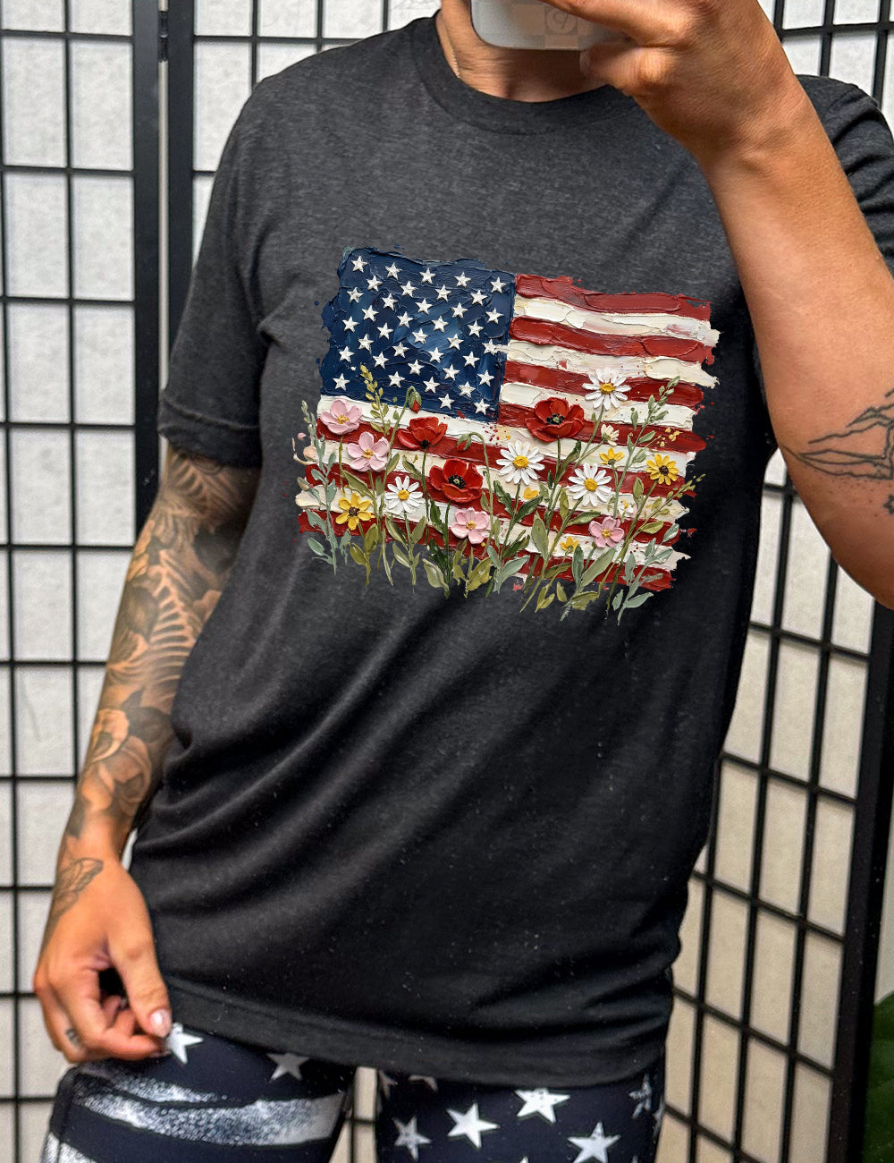 black unisex tee with the graphic of an American flag with flowers in front of it, with the style of the graphic looking made of oil paint 