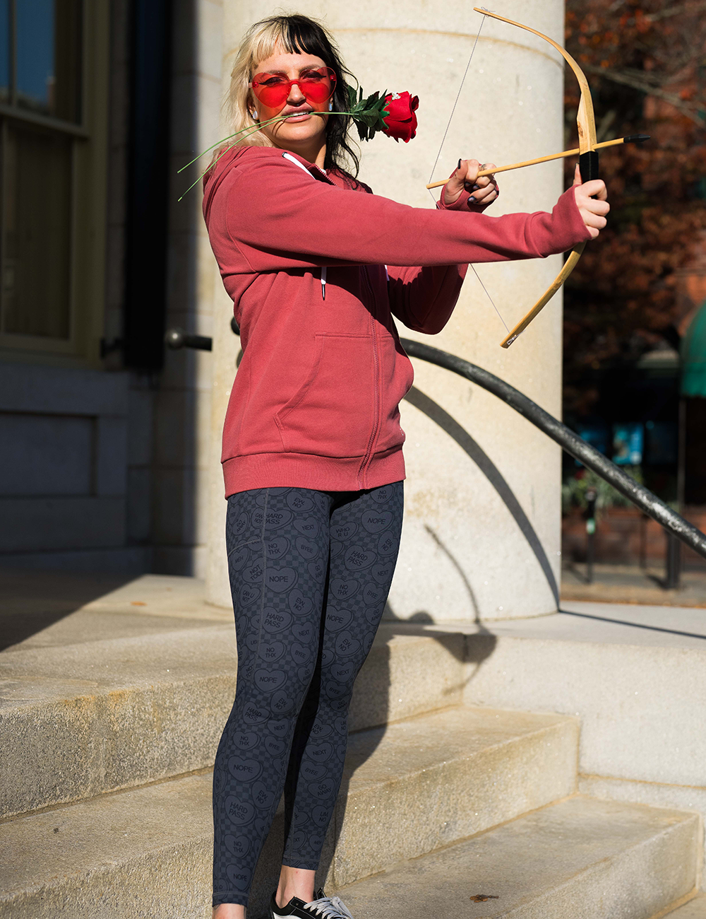  black and grey heart patterned leggings with a red sweatshirt, holding cupid's bow and arrow