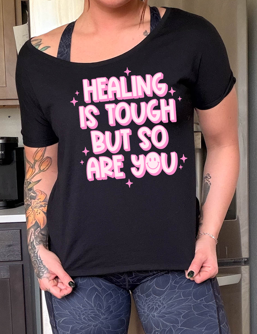 black slouchy tee with the words "healing is tough but so are you" on it, with a smiley face in the word "you", and little star graphics scattered around