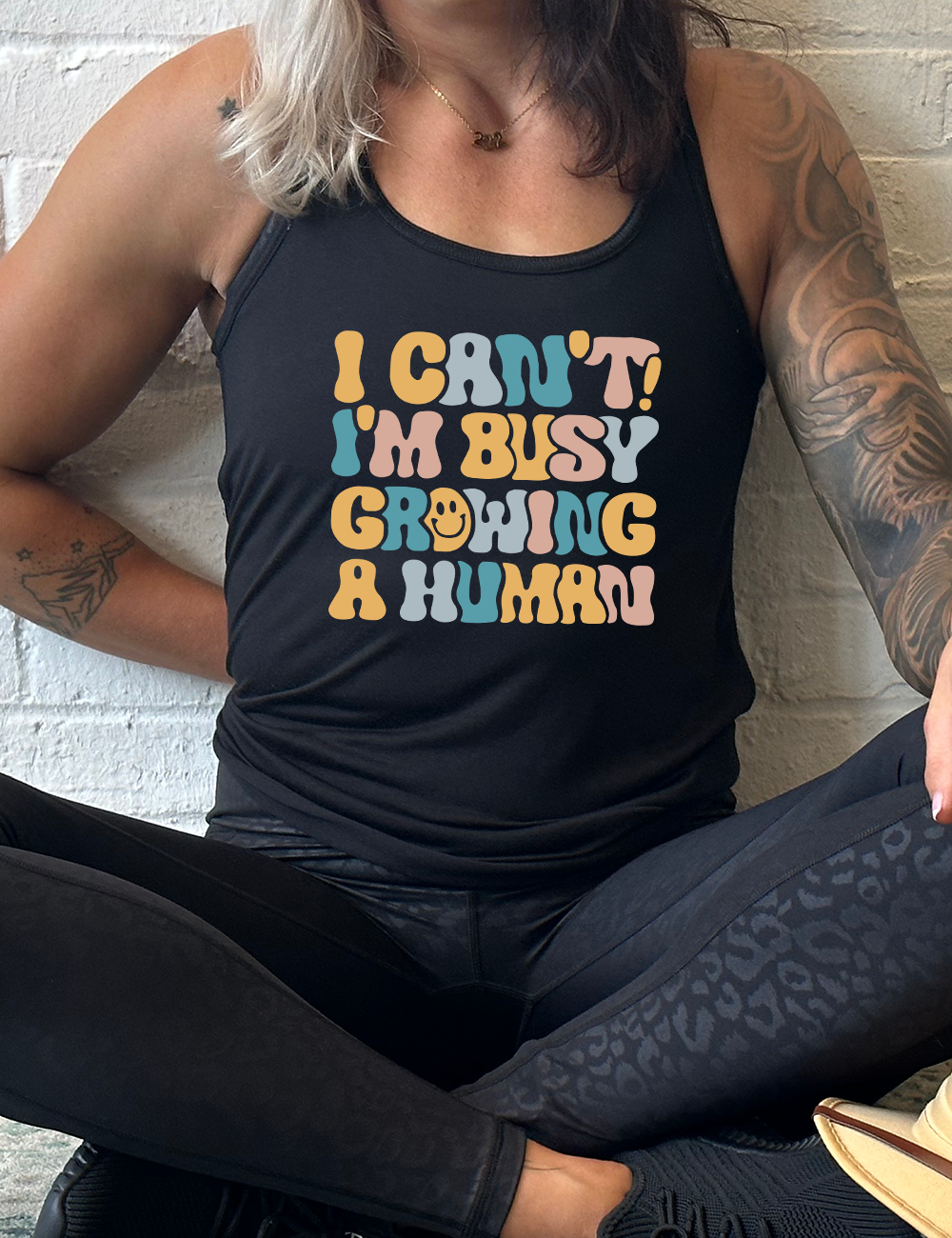 black racerback flowy tank with the words "I can't! I'm busy growing a human" on it in colorful font, with a smiley face in the "o" of the word "growing"