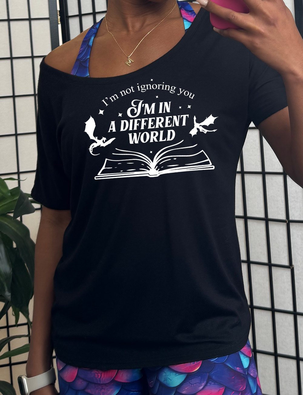black slouchy tee with the words "I'm not ignoring you I'm in a different world" on it, with the graphic of a large book, lying open, and two little dragons flying next to the words, a few stars scattered around