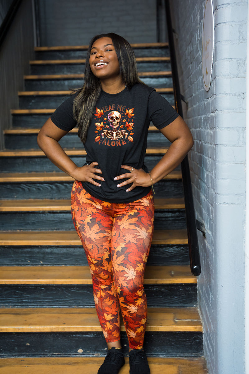 A woman posing on an indoor staircase wearing a black graphic t-shirt and orange/red autumn leaf patterned leggings.