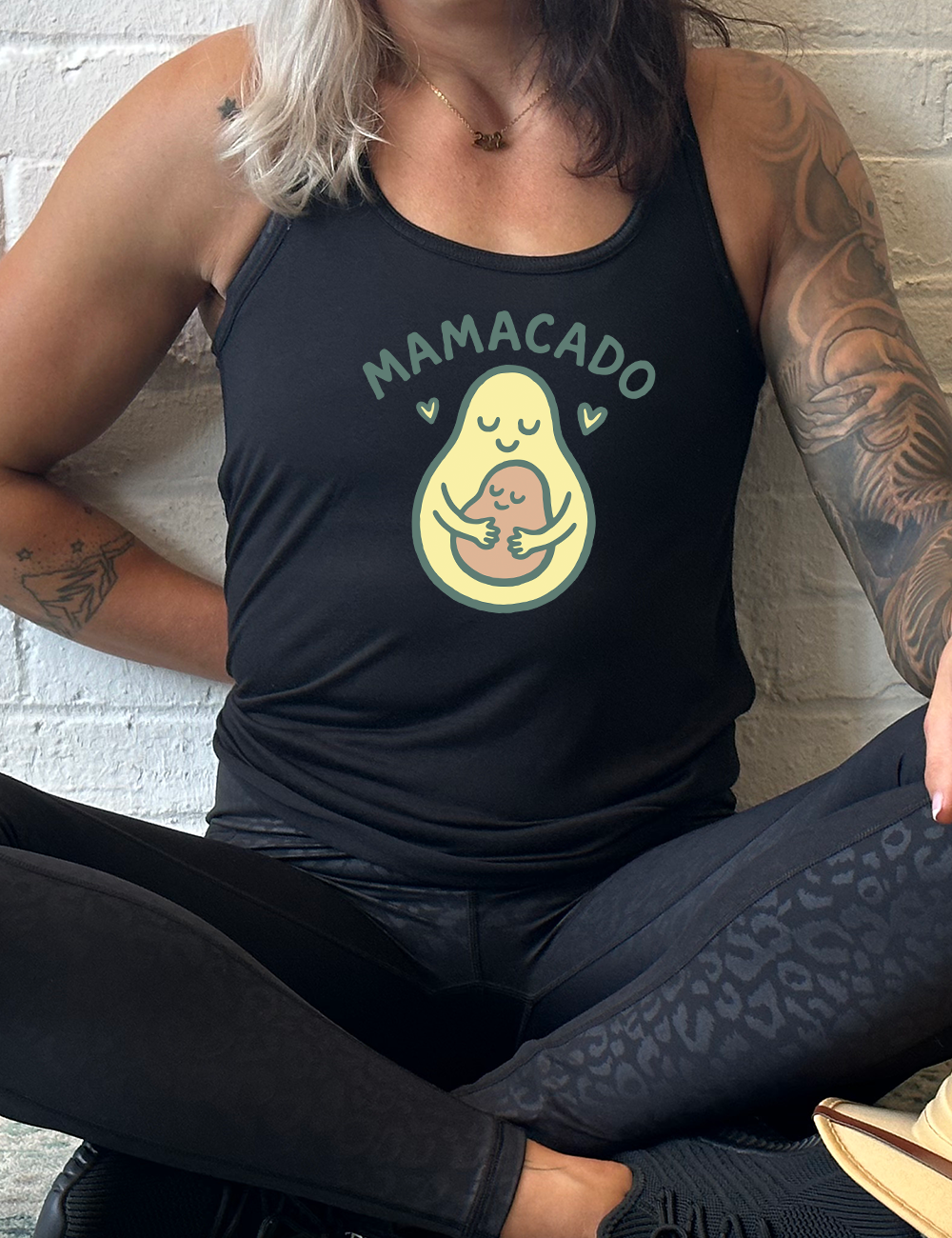 black racerback flowy tank with the words "mamacado" on it, with the graphic of an avocado, peacefully smiling, with arms, holding her smiling baby (the color and placement of the avocado pit - but in the shape of an avocado), with a little heart on either side 