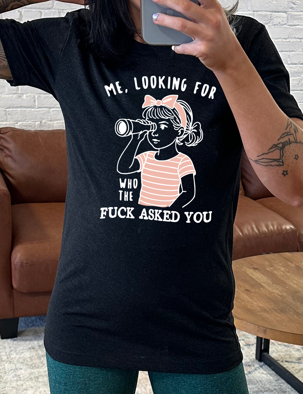 black unisex tee with the graphic of a young girl, wearing a striped pink shirt and a pink bowed headband and looking through a telescope, and the words "me, looking for who the fuck asked you" on it
