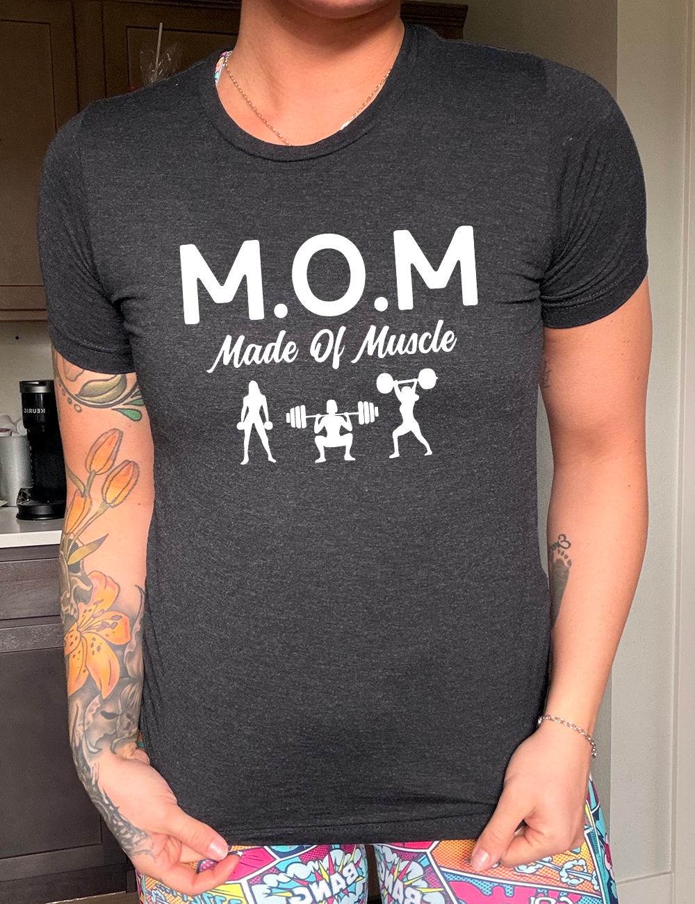 black unisex tee with the word "mom" in large letters as an acronym for the smaller words below it saying "made of muscle", and the graphic of three female silhouettes holding weights