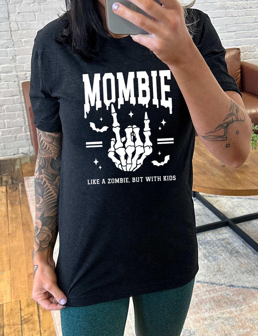 black unisex tee with the big word "mombie" on it, with the graphic of a skeletal hand giving the rocker sign, with a couple bats and a number of little stars, and two equal signs, with small words underneath it all saying "like a zombie, but with kids"