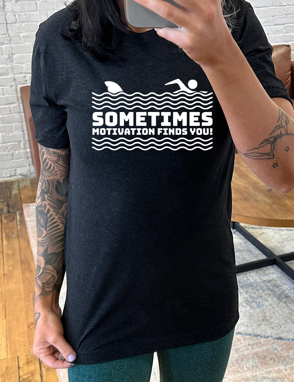 black unisex tee with the words "sometimes motivation finds you!" in between the lined graphic of waves of the sea, and on top of the waves is the graphic of a figure swimming away from a fin in the water behind