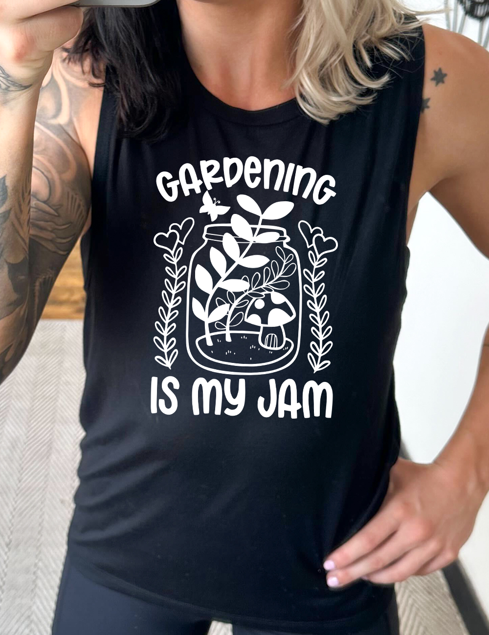 black muscle tank with the words "gardening is my jam" on it, and the graphic of a jar with plants and a mushroom in it, a little butterfly flying above it, and a plant (with hearts for flowers on them) on both sides of it