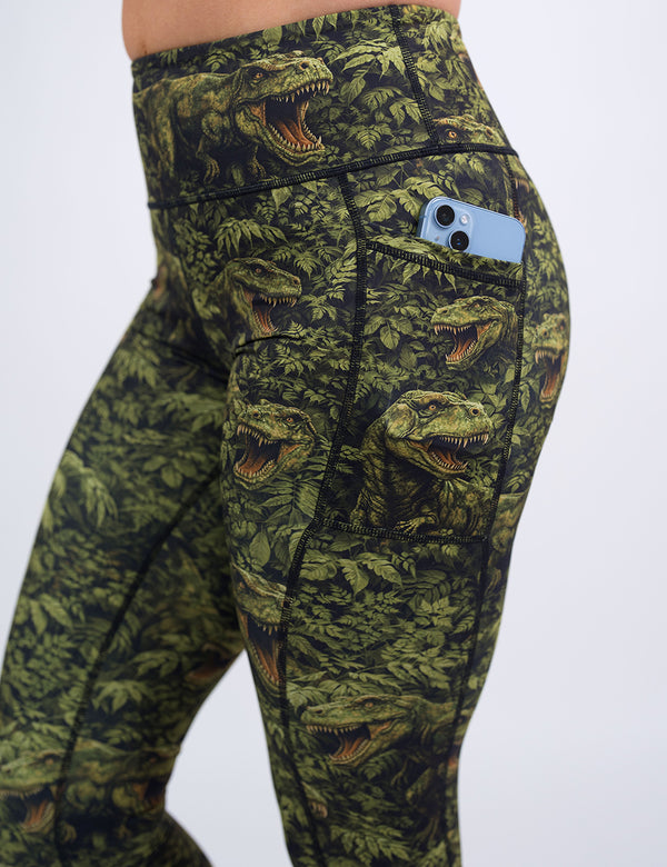 close up of the green t-rex in a forest leggings holding a phone in its pocket against a white background